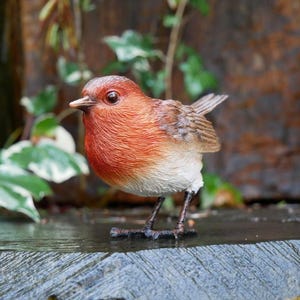 May include: A detailed figurine of a robin bird with a red breast, brown wings, and a white belly. The bird is standing on a wooden surface, with greenery in the background. The bird's beak is open, and its eyes are visible.