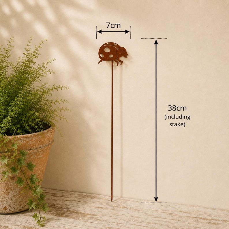 May include: A brown metal garden stake featuring a ladybug silhouette at the top. The ladybug ornament measures 7 cm in width, and the total height of the stake including the pointed end is 38 cm. The design is a simple, flat metal cutout.