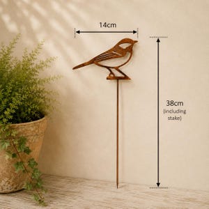May include: A rust-colored metal bird garden stake with a cutout design. The bird figure measures 14 cm in width, and the total height of the stake including the bird is 38 cm. The object features a thin, straight metal rod for ground insertion.