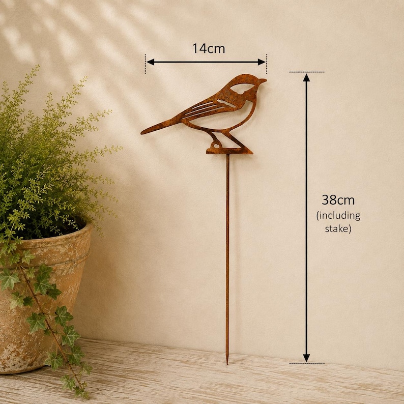 May include: A rust-colored metal bird garden stake with a cutout design. The bird figure measures 14 cm in width, and the total height of the stake including the bird is 38 cm. The object features a thin, straight metal rod for ground insertion.