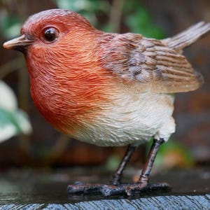 May include: A detailed figurine of a European robin. The bird has a red-orange breast, a brown back, and a white belly. Its small, dark legs are visible, and it is perched on a surface. The figurine is a decorative item.