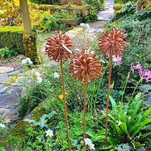 May include: Three brown metal flower sculptures are displayed in a garden. The flowers have a layered, textured design and are mounted on slender metal stems. The garden setting includes lush greenery, stone pathways, and various flowering plants.