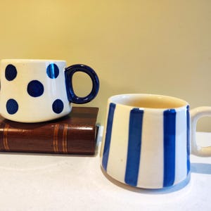 May include: Two ceramic mugs. One is white with blue polka dots and a blue handle. The other is white with blue vertical stripes and a white handle. Both mugs are on a brown book.