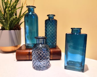 Könnte beinhalten: Vier türkisfarbene Glasvasen in verschiedenen Formen und Texturen. Eine ist eine hohe, zylindrische Flasche, eine weitere eine quadratische Flasche, und zwei sind kleinere, dekorative Flaschen. Die Vasen sind auf einem Holzbuch angeordnet.