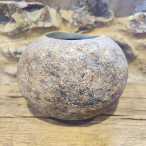 May include: A spherical, textured stone vase with a wide opening. The vase has a mottled appearance with shades of brown, tan, and grey. It sits on a wooden surface, with a stone wall in the background. This decorative vase is suitable for home decor.