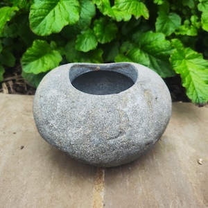 May include: A grey, rounded stone planter with a wide opening. The stone has a textured surface and sits on a light brown stone surface. Green leafy plants are in the background.