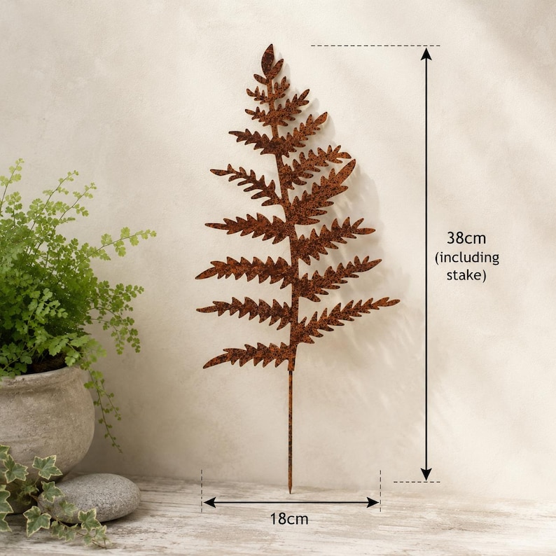 May include: A rust-colored metal fern leaf garden stake with a textured finish. The decorative plant silhouette features a pointed tip and a thin stem base. The total height including the stake is 38cm, and the width of the fern leaf is 18cm.