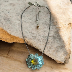 May include: A silver-toned necklace featuring a flower pendant. The pendant has a yellow center surrounded by light blue petals. The necklace chain is dark gray and rests on a light brown surface.