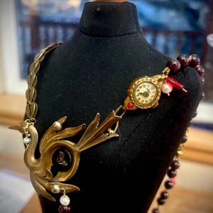 May include: A unique, handcrafted necklace featuring a vintage watch face, a brass bird pendant, and a strand of dark red beads. The necklace has a steampunk aesthetic, with intricate details and a vintage charm.