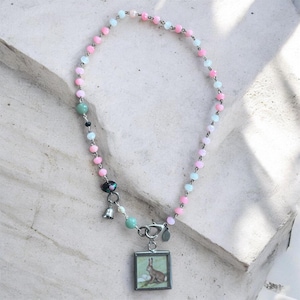 Op de afbeelding: Een ketting met een hanger met een konijnenillustratie. De ketting heeft een kralenketting in pastelroze en blauw. Extra bedels zijn een kleine bel en een decoratieve kralencluster. De hanger is vierkant en zilverkleurig omlijst.