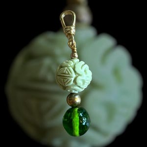May include: A pendant with a gold-colored clasp and wire. The pendant features a carved, pale green sphere with geometric designs, a small gold bead, and a dark green, striped glass bead. The background is blurred.