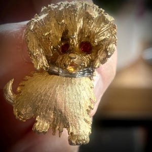 May include: A gold-toned metal dog brooch with red rhinestone eyes and a textured finish. The dog has a collar and a fluffy appearance, with detailed fur-like texturing throughout its body. A vintage-style accessory.