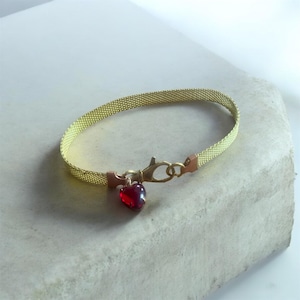 Puede incluir: Una pulsera de malla dorada con un dije en forma de corazón rojo. La pulsera tiene un cierre de broche y pequeños detalles de color cobre. El dije es de color rojo intenso y parece estar hecho de vidrio o un material similar.