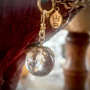 May include: A clear, spherical pendant with gold-colored accents and a small tag with a crown design and the words "Stories Retold." The pendant is attached to a gold-colored chain and clasp. The background is a blurred maroon color.