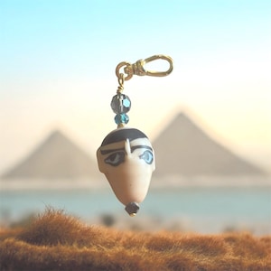 May include: A gold-colored charm with an Egyptian pharaoh design. The charm features a cream-colored face with black and blue details, a black headdress, and blue eyes. It hangs from a gold-colored clasp and is adorned with blue and gray beads.