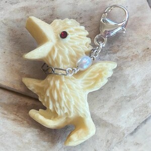 Op de afbeelding: Een crèmekleurige vogelbedel met een rode edelsteenoog en een zilveren kettingkraag. De vogel heeft uitgespreide vleugels en is bevestigd aan een zilveren sluiting. Een kleine, iriserende parel hangt aan de ketting.