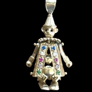 Puede incluir: Colgante dorado con forma de payaso. El payaso tiene un sombrero en forma de cono, un cuello con volantes y un abrigo adornado con piedras preciosas de colores. El colgante cuelga de un pequeño lazo en la parte superior.