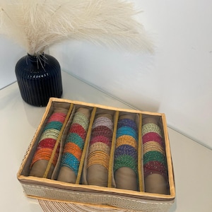 May include: A beige fabric bangle box with a clear plastic lid. The box has six compartments filled with colourful glass bangles. The bangles are arranged in rows and are of different colours, including red, orange, yellow, green, blue, and purple.