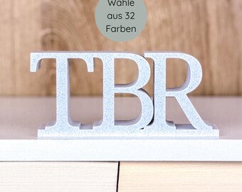 May include: A light gray TBR bookend set on a white shelf, with the letters standing upright. A circular graphic above the letters reads "Wähle aus 32 Farben". The background is a light brown wooden surface.