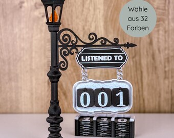 May include: A black decorative street lamp with a sign that reads "LISTENED TO" and a counter displaying "001". The sign is hanging from a decorative arm. The lamp has an orange light. The text "Wähle aus 32 Farben" is in a circle.