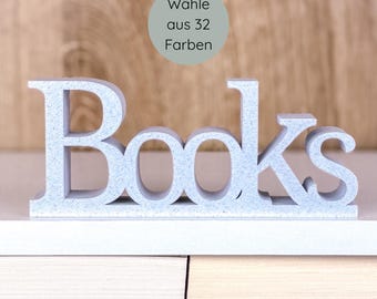 May include: A light blue, freestanding "Books" sign. The word is in a bold, serif font. A circular graphic in the background reads "Wähle aus 32 Farben". The sign is displayed on a white shelf against a wooden background.