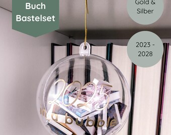 May include: A clear spherical ornament filled with miniature books, featuring gold text that reads 2025 in a bubble. The ornament hangs from a gold-colored metal hook attached to a white ceiling mount, showcasing a DIY mini-book craft kit design.