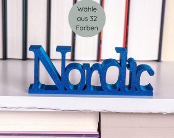 May include: A blue, three-dimensional "Nordic" word sign. The sign is displayed on a white shelf in front of a stack of books. A gray circle with the text "Wähle aus 32 Farben" is visible.