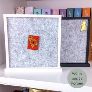 Könnte beinhalten: Zwei gerahmte graue Filztafeln, eine weiße und eine schwarze. Der weiße Rahmen zeigt eine rot-orangefarbene Anstecknadel mit einem Drachenaugen-Design. Der schwarze Rahmen hat eine goldene Anstecknadel. Der Text lautet "Wählen Sie aus 32 Farben."