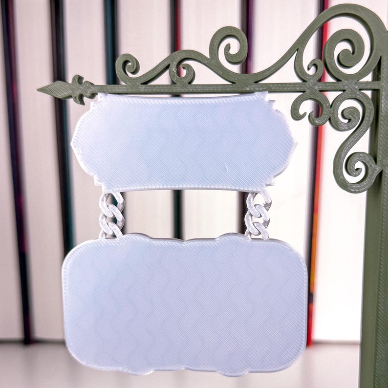 May include: An olive green sign with two light blue, oval-shaped panels. The panels are suspended by chain links from a decorative, scrolled top. The sign has an ornate, vintage design, suitable for a shop or business.