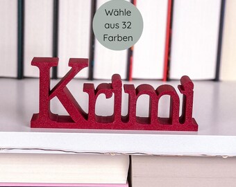 May include: A red wooden cutout of the word "Krimi" is displayed on a shelf. The word is in a bold, sans-serif font. A circular graphic in the background reads "Wähle aus 32 Farben". The shelf is positioned in front of a blurred background of books.