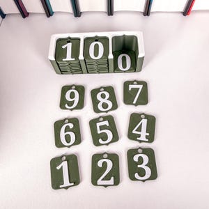 May include: A collection of green and white number tags, featuring numbers 1 to 10. The white numbers are on green square tags. A white and green holder displays the number 100. The tags have a small hole at the top.