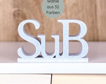May include: A light blue, freestanding decorative sign with the letters "SuB" in a stylized font. The sign is on a white surface, with the text "Wähle aus 32 Farben" in a circular graphic in the background.