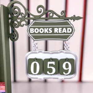 May include: A green and white "BOOKS READ" sign with a counter showing "059". The sign features an ornate, scrollwork bracket. The counter is designed to track the number of books read.