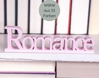 May include: A light pink, freestanding "Romance" word sign, displayed on a white shelf in front of a backdrop of books. A circular graphic above the sign reads "Wähle aus 32 Farben". The sign is a decorative item.