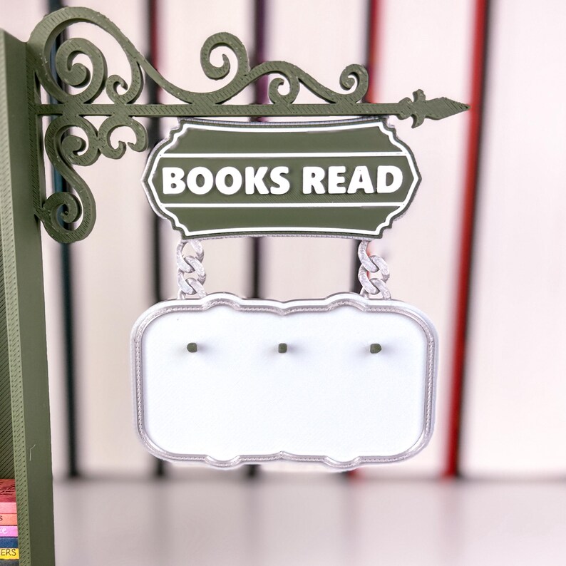 May include: A green metal sign with the words "BOOKS READ" in white. The sign features a decorative scroll design and a blank, white lower section. A blurred collection of books forms the background.