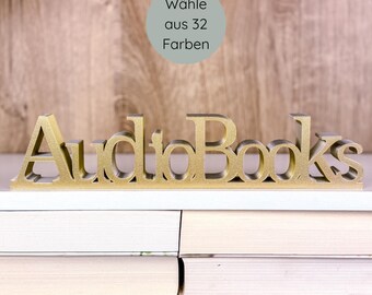 May include: A gold-colored "AudioBooks" sign sits atop a stack of books. The sign is in a serif font and is placed on a white shelf. A light green circle with German text is in the background.