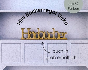 May include: A gold-colored, decorative word "Hörbücher" sits on a shelf within a light gray cabinet. The cabinet has a simple design with three sections. Text above reads "Mini Bücherregal Deko" and "Wähle aus 32 Farben".