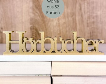 May include: A gold-colored wooden sign that reads "Hörbücher" in stylized lettering. The sign is displayed on a white surface, with a stack of books in the foreground. A circular graphic above the sign says "Wähle aus 32 Farben".