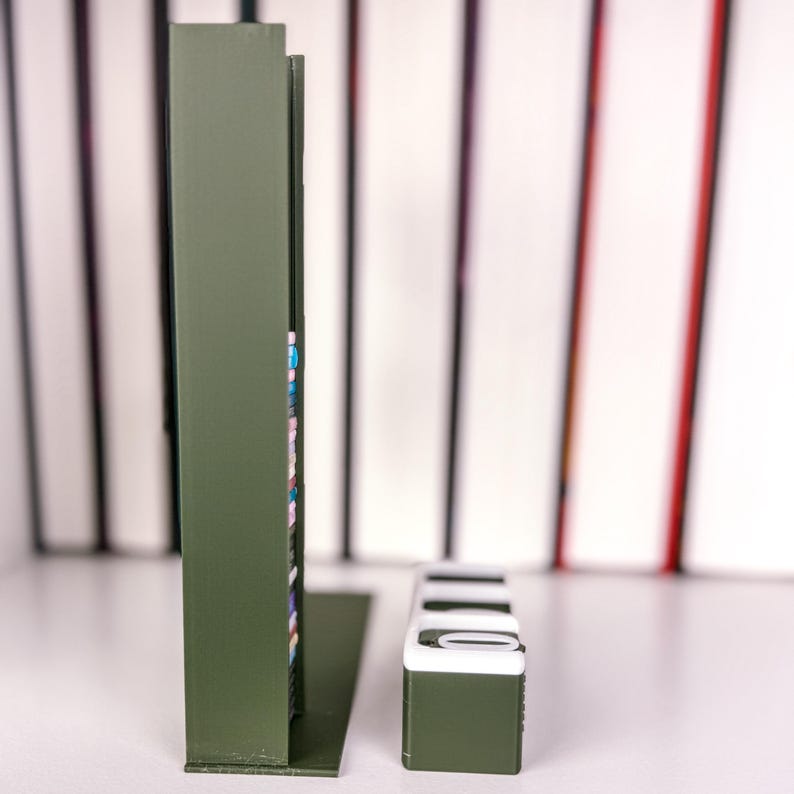 May include: Olive green metal bookends with a rectangular shape and a small, matching accessory. The bookends have a vertical design, and the accessory has a white top. The background features a blurred array of books.