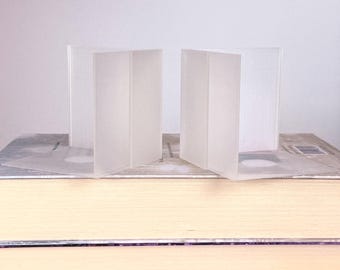 May include: Two frosted glass bookends stand on a stack of books. The bookends are rectangular and have a folded design. The books have yellow and purple covers. The background is a plain white.