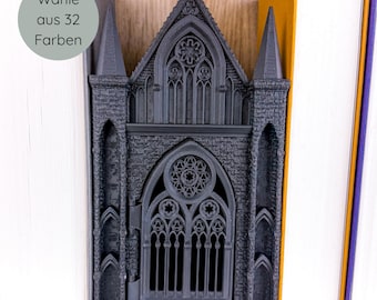 May include: A dark gray, gothic-style bookend shaped like a cathedral. The bookend features intricate architectural details, including arched windows and spires. The text "Wähle aus 32 Farben" is visible on the left side. The bookend is positioned between two books.