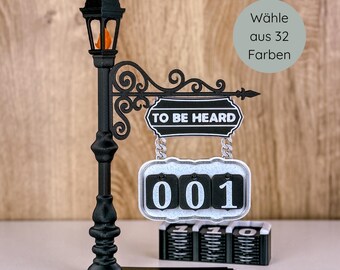 May include: A black decorative street lamp with a sign that reads "TO BE HEARD" and a counter displaying "001". The lamp has an orange flame and a base with a small platform. The sign has a decorative scroll design.