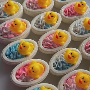 May include: A collection of oval-shaped bath bombs, each featuring a small yellow rubber duck figurine resting atop a swirl of pink or blue textured cream. The bath bombs have a white, firm base and are arranged in a repeating pattern.