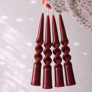 May include: Four burgundy-colored spiral taper candles held by a person. The candles have a twisted design and are suspended by white strings. The background is a light, neutral color.