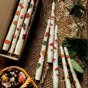 May include: A collection of white taper candles decorated with small, raised wax appliques of orange pumpkins, red and white mushrooms, and colorful autumn leaves. Some candles are inside a cardboard box, while others are arranged nearby.