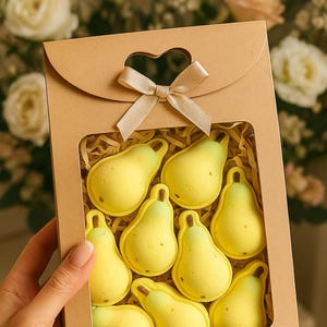 May include: A gift box with a heart-shaped window and a tied bow contains eight yellow pear-shaped treats. The pears have a light green top and brown speckles. The box is held by a hand.