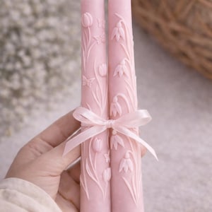 May include: Two pink pillar candles tied with a pink ribbon. The candles are decorated with embossed floral and butterfly designs. The candles are approximately 8 inches tall.