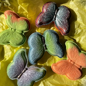 May include: An assortment of colorful butterfly-shaped bath bombs. The bath bombs are in shades of green, orange, purple, and blue, with a shimmering, textured finish. They rest on a bed of yellow tissue paper.