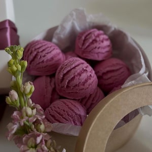 May include: A collection of pink, scoop-shaped bath bombs nestled in a round, open cardboard container lined with white tissue paper. A sprig of pink and green flowers is in the foreground, with a white box and purple ribbon in the background.