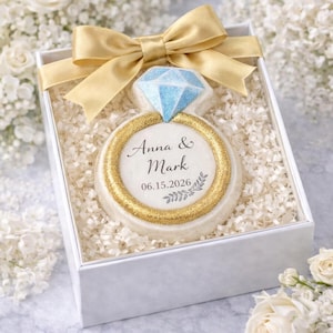 May include: A ring-shaped cookie in a white box, decorated with a gold glitter band and a blue diamond. The cookie is inscribed with "Anna & Mark" and the date 06.15.2026. A gold ribbon bow adorns the box, surrounded by white flowers.